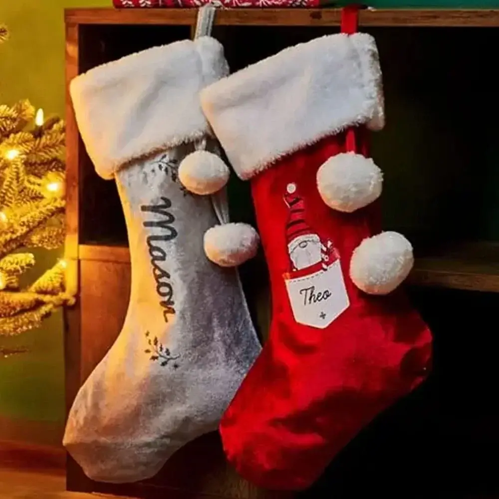 Red velvet Christmas gift sack with white trim and pom-pom drawstring, featuring a festive Santa and reindeer design with the name Annabelle, placed beside wrapped presents and a stocking near a decorated Christmas tree.